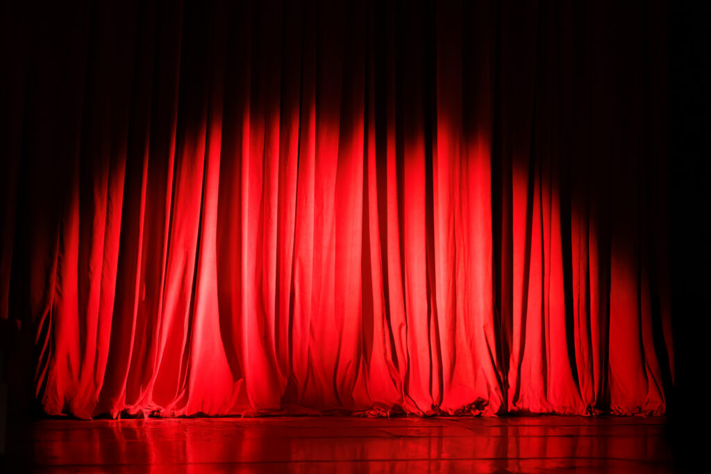 
A dramatic image of closed red velvet curtains on a theater stage, lit from the front by two spotlights. The curtains are slightly wrinkled and bunched at the bottom, and the light reflects on the polished wooden floor in front of them.