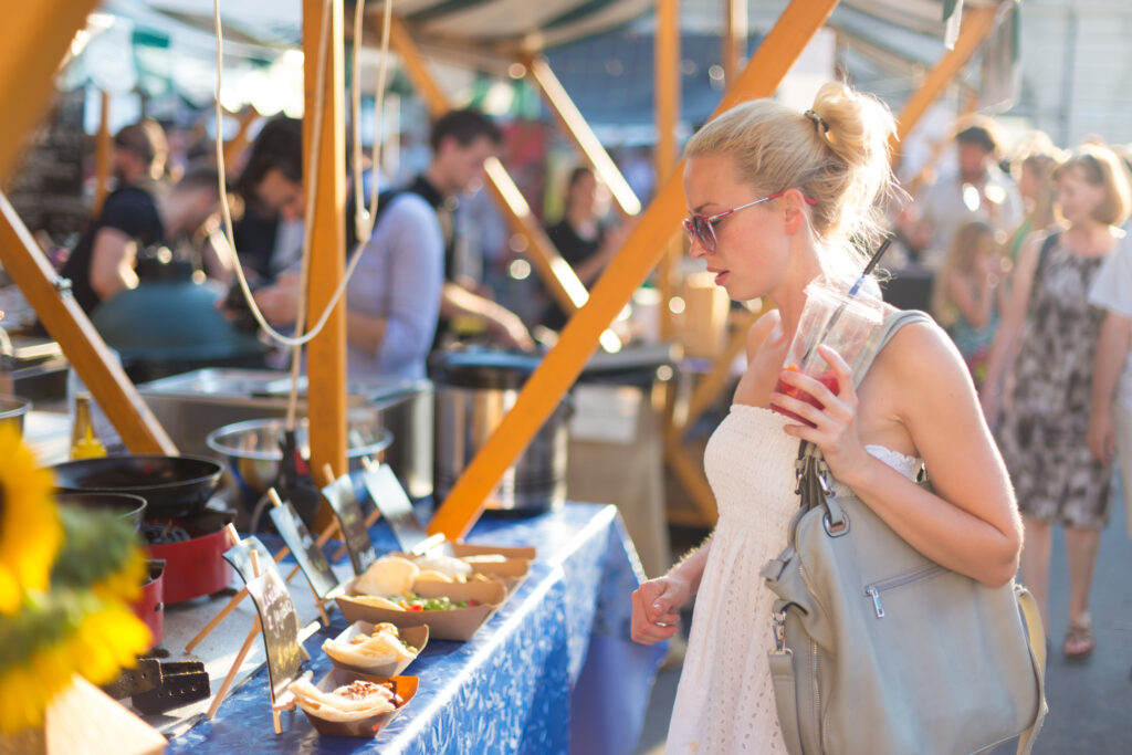 A blonde woman with her hair in a bun and wearing sunglasses and a white dress is looking at food samples on a table at an outdoor market. She holds a plastic cup with a red drink in her left hand and a gray purse on her shoulder. The background is filled with blurred people and various food booths.