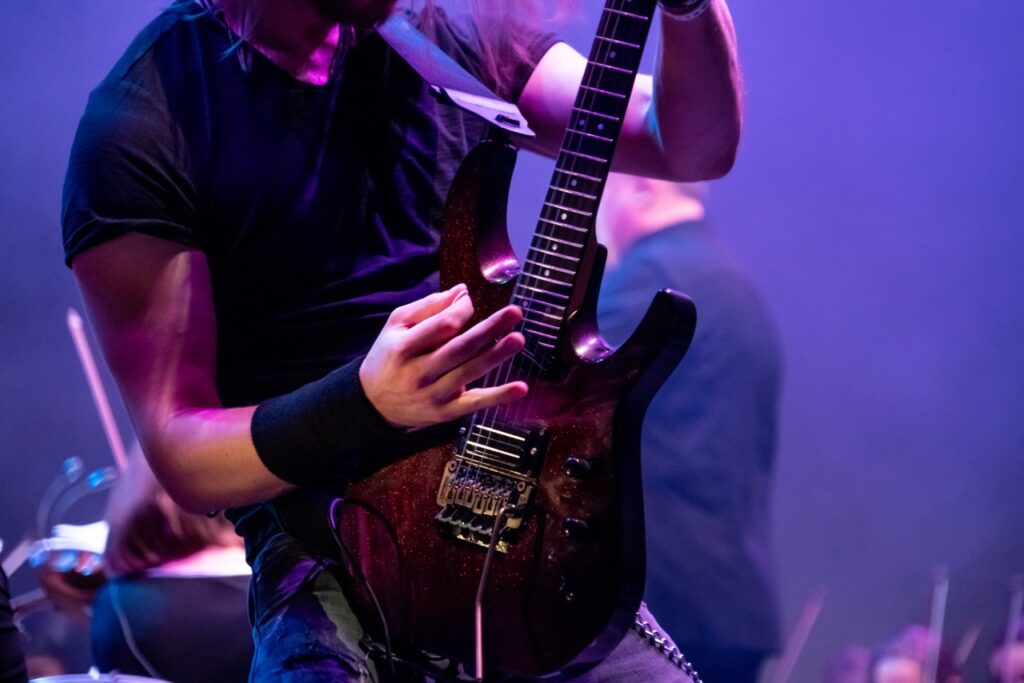 A person in a black t-shirt and wristband is playing an electric guitar with a dark red, sparkly finish. The stage is lit with vibrant purple and blue lights. Another musician is visible in the blurred background.