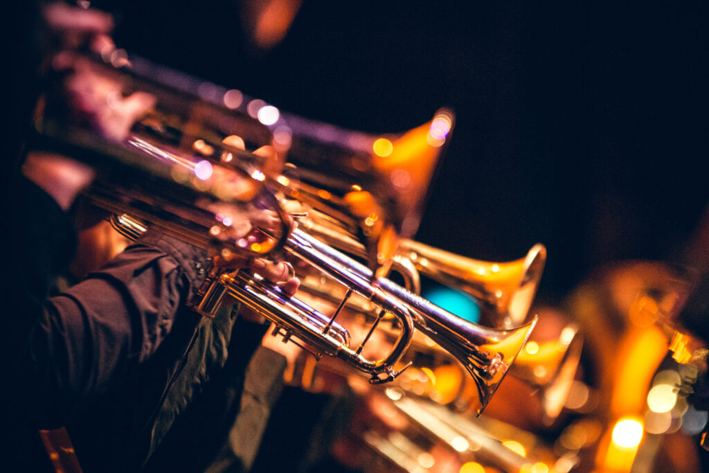 A row of trumpet players are performing on a dimly lit stage. The brass instruments are shiny and reflect the warm, golden stage lights. The focus is on the middle trumpet, while the others are slightly blurred in the foreground and background.