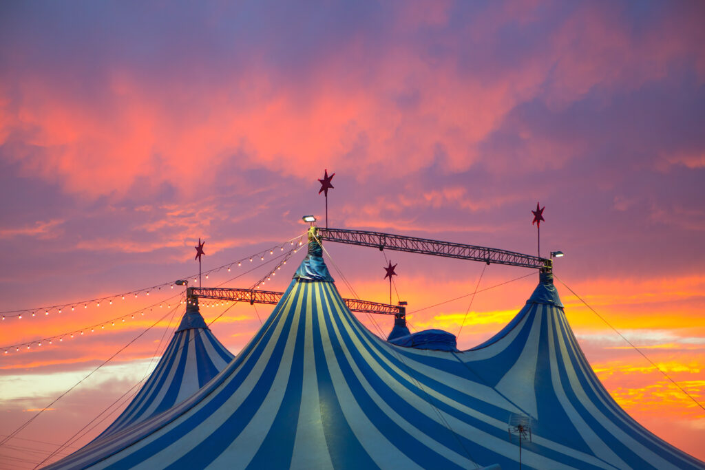 A vibrant, blue-and-white striped circus tent stands against a dramatic sunset sky filled with hues of orange, pink, and purple clouds. Three red star ornaments top the peaks of the tent.