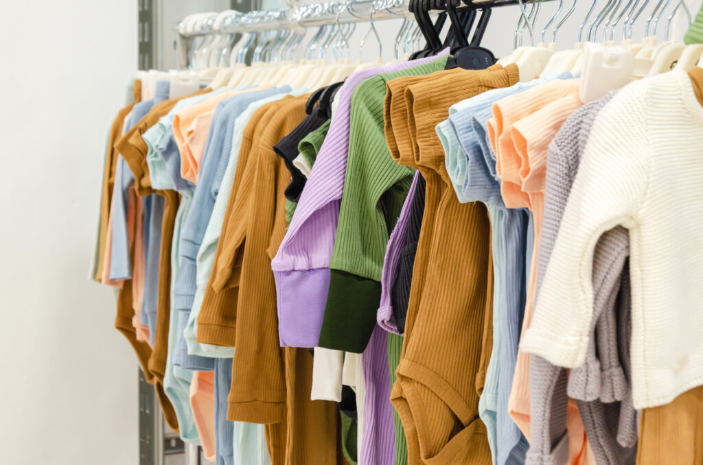 A rack of baby or children's clothing is on display in a store. The clothes are in a variety of soft, muted colors like brown, olive green, lavender, blue, and cream.