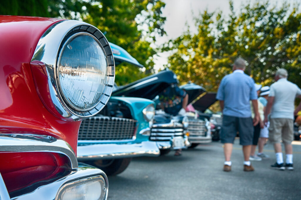 A classic car show on a sunny day. The front of a red vintage car is in sharp focus, with its chrome bumper and shiny round headlight. The background is blurred, showing a line of other classic cars with their hoods open and people walking around.