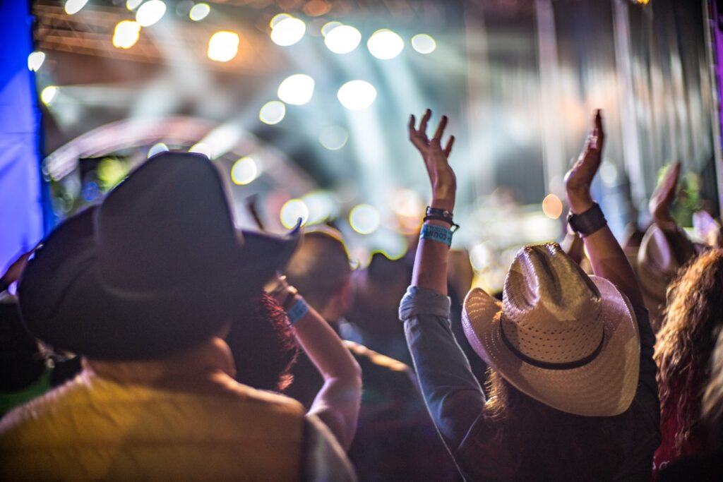 A blurred, low-angle shot of a crowd at a concert, with a person in a dark cowboy hat on the left and a person in a light-colored cowboy hat on the right with their hands raised in the air. Bright, out-of-focus stage lights are visible in the background.