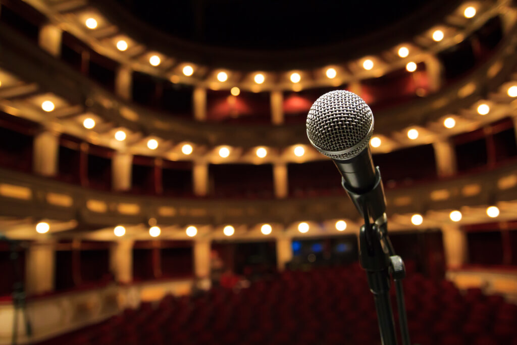 A close-up of a microphone on a stand, placed on a stage. The background is a blurred view of a beautiful theater with red seats and glowing yellow lights lining the balconies.