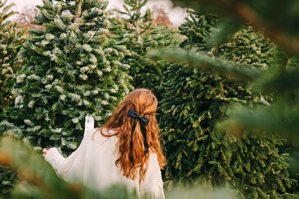 A woman with long hair and a black bow is seen from behind as she chooses a Christmas tree at a farm.