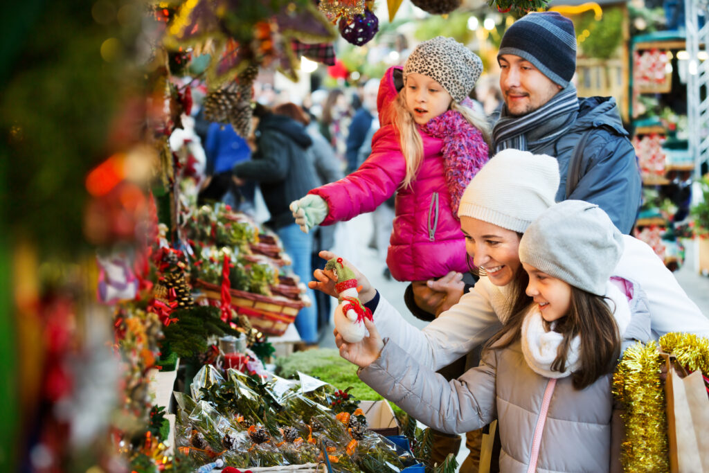 Ordinary family with little girls standing at coniferous Christmas souvenirs counter. Focus on women
