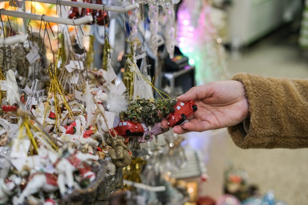 Close-up of female hand in brown fluffy jacket selecting red vintage car Christmas ornament from crowded retail display rack. Holiday shopping, choosing decorations, Christmas fair