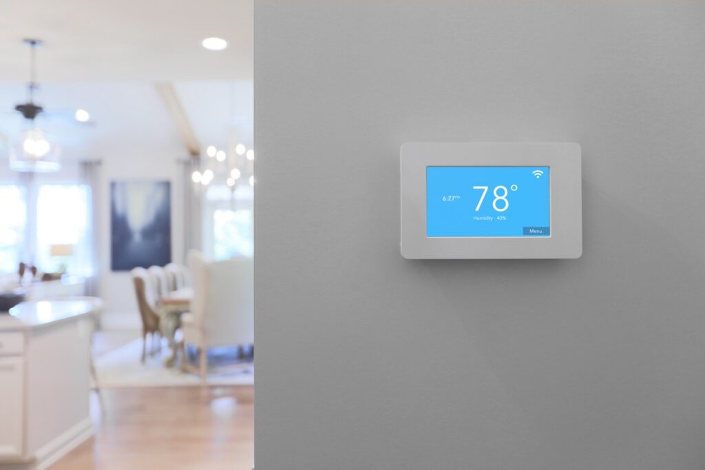 Modern smart thermostat mounted on a clean grey interior wall with an open-concept kitchen and living area blurred in the background, illustrating energy efficiency upgrades for home buyers in Greenville SC.