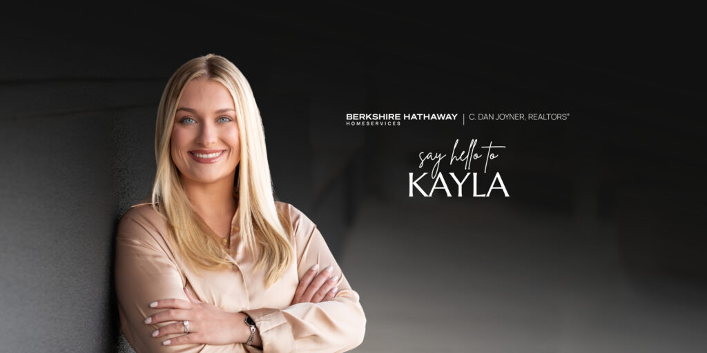 A wide horizontal banner graphic. On the left, REALTOR® Kayla Guin smiles with crossed arms in a champagne-colored blouse against a textured dark grey wall. The right side has a blurred dark background with the Berkshire Hathaway HomeServices C. Dan Joyner, REALTORS® logo. Stylized white text reads: 'say hello to KAYLA' with a small curved arrow pointing to her.