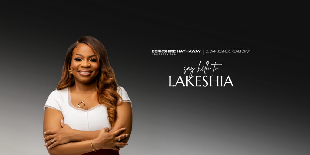 A wide horizontal website banner featuring REALTOR® Lakeshia Glenn smiling on the left in a white top with arms crossed against a dark gray background. On the right, white text reads 'say hello to LAKESHIA' beneath the Berkshire Hathaway HomeServices C. Dan Joyner, REALTORS® logo
