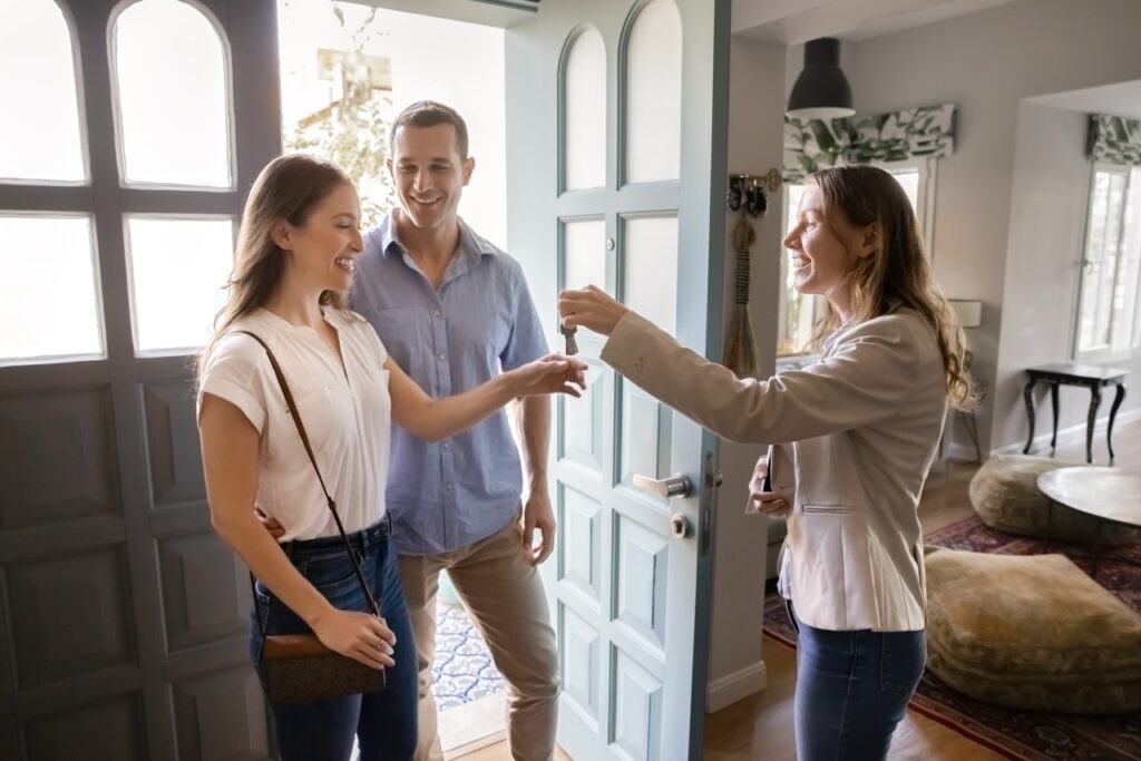 A smiling real estate agent handing house keys to a happy couple at the front door on closing day after finalizing settlement costs.