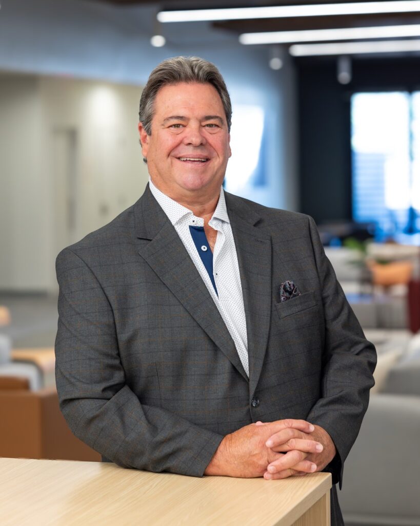 A professional headshot of Bill Hentges, a real estate agent with Berkshire Hathaway HomeServices C. Dan Joyner, REALTORS®, at the Woodruff at Five Forks office in Greenville/Simpsonville. He is a smiling middle-aged man with grey-brown hair, wearing a dark grey plaid blazer, a patterned white collared shirt with blue trim, and a pocket square, posing with his hands clasped over a light wood railing in a modern, naturally lit office environment.