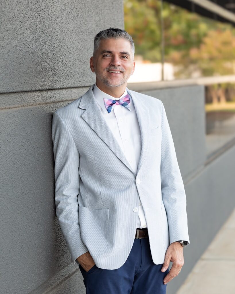 A professional headshot of REALTOR® Jorge Lopez, a bilingual agent with Ronda Holder & Associates at the Woodruff at Five Forks office of Berkshire Hathaway HomeServices C. Dan Joyner, REALTORS®, smiling in business-casual attire against a neutral studio background.